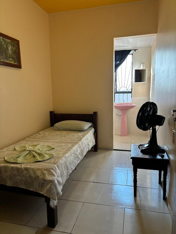 A bedroom with a bed, a bedside table with a fan, a pink sink, and a window with curtains. - Hotel São Luis (Pinhao)