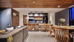 A modern living room with a sofa, dining table, and chairs. There is a kitchen area with cabinets and a refrigerator.