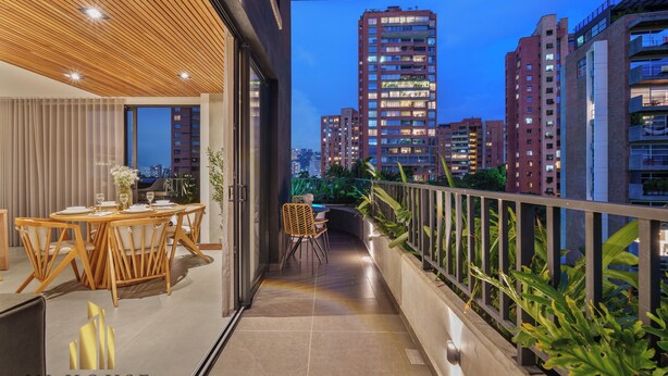 A balcony with a dining area, potted plants, and a cityscape view at dusk.