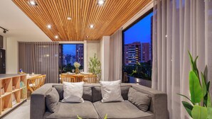 A modern living room with a grey sectional sofa, a wooden dining table, and a city view through large windows.