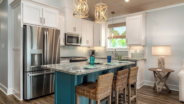 Una cocina moderna con armarios blancos, electrodomésticos de acero inoxidable, una isla verde azulada con taburetes y luces colgantes.