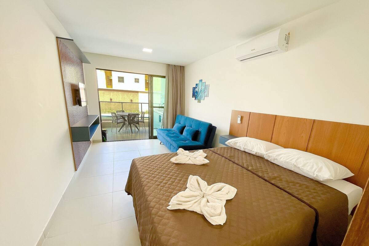 A hotel room with a bed, a blue sofa, a balcony with a table and chairs, and an air conditioning unit.