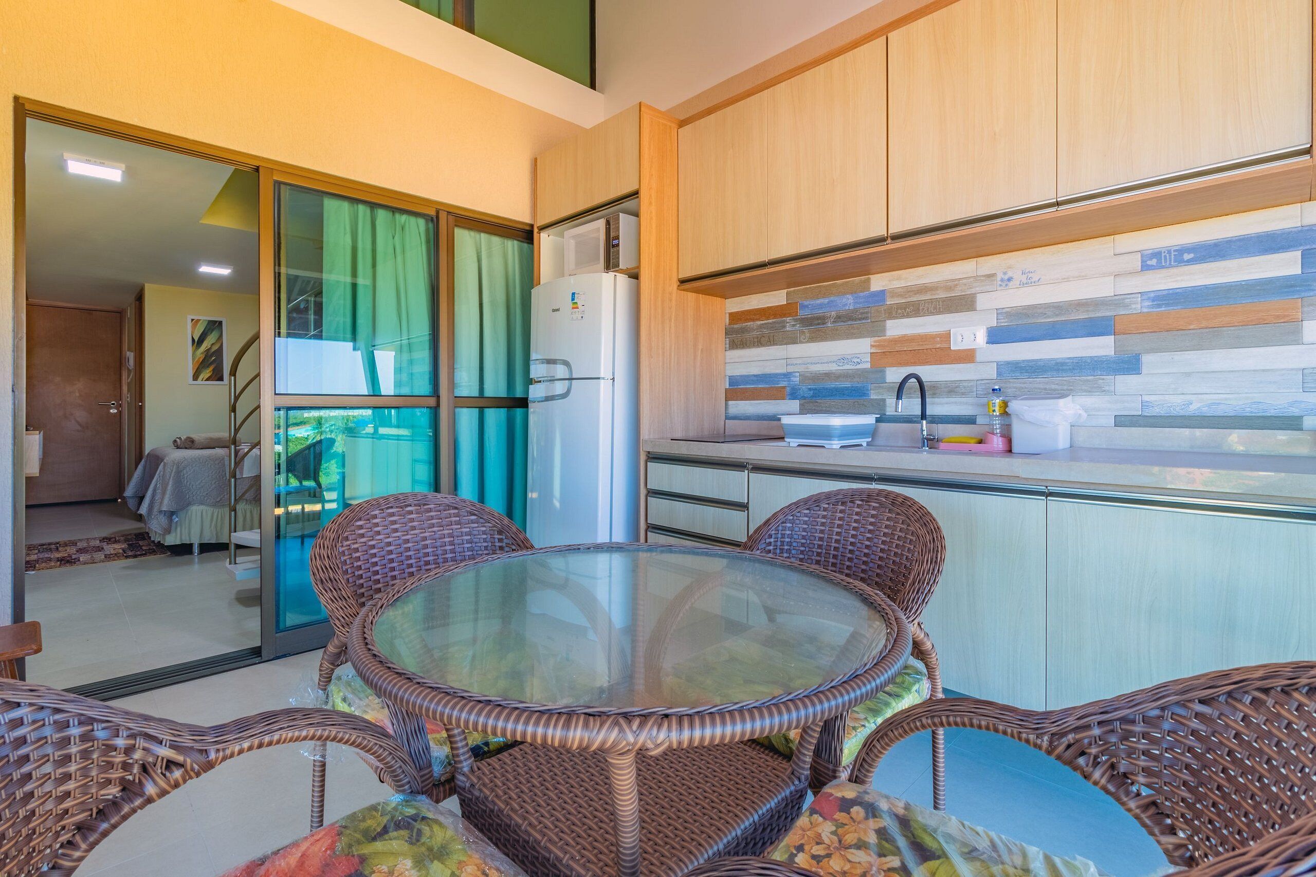 A modern kitchen with a glass-top dining table and wicker chairs, a refrigerator, and a view of a balcony.