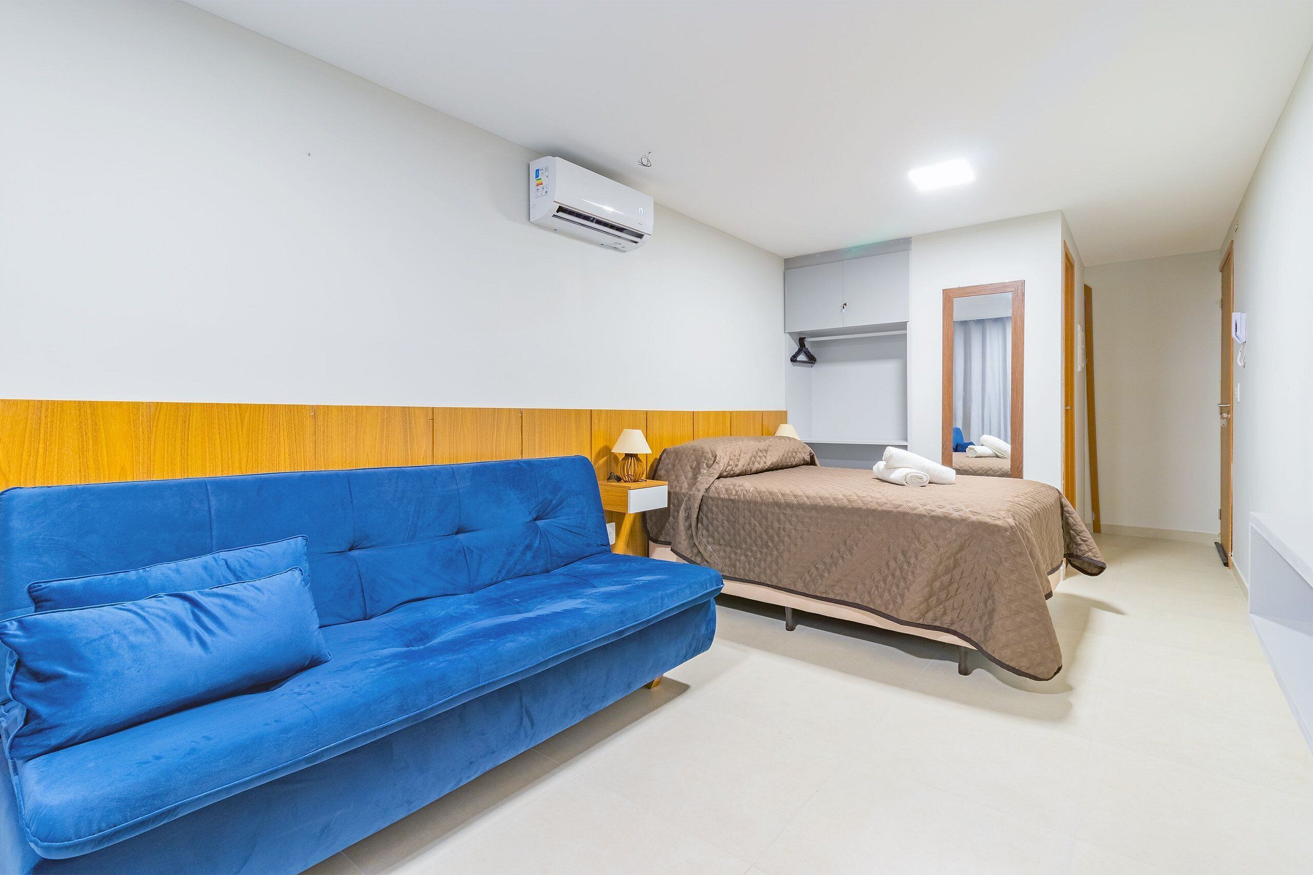 Ein Zimmer mit einem blauen Sofa, einem Bett mit brauner Bettdecke, einem kleinen Tisch mit einer Lampe und einer Klimaanlage an der Wand.