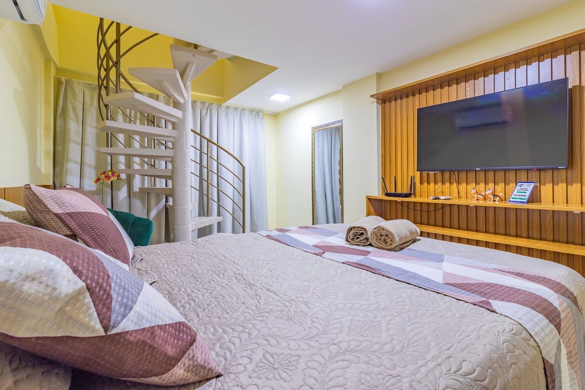 A hotel room with a bed, a TV, a wooden panel wall, a window with curtains, and a spiral staircase.