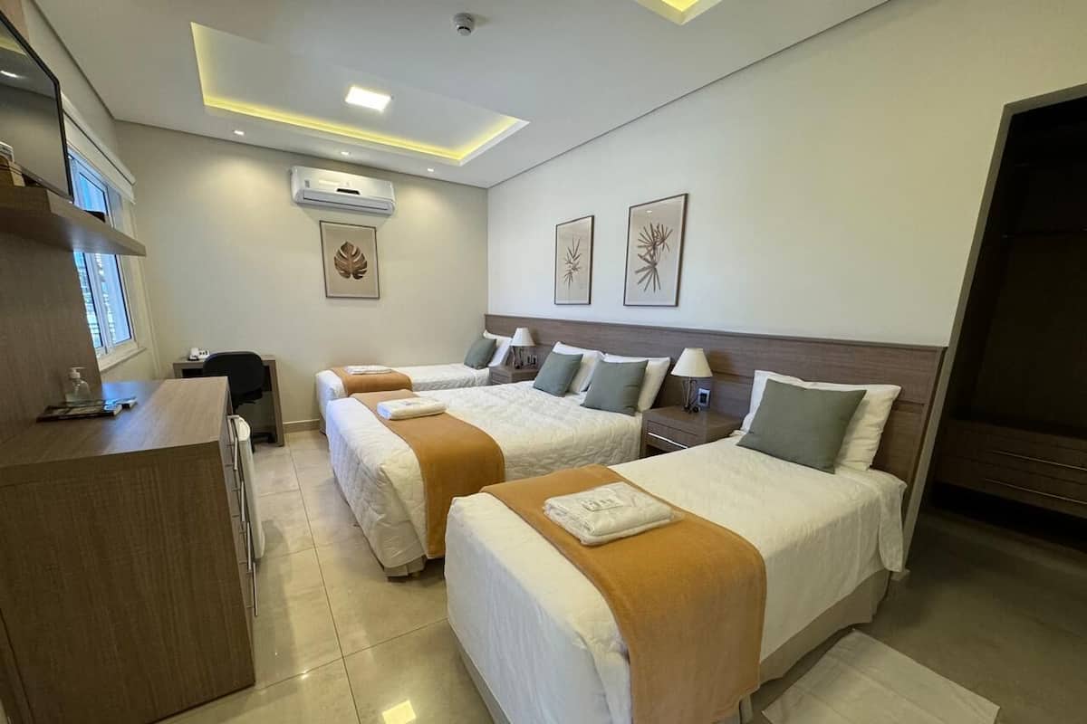A hotel room with two beds, a desk, and a chair. There are wall-mounted air conditioning units and framed artwork above the beds.