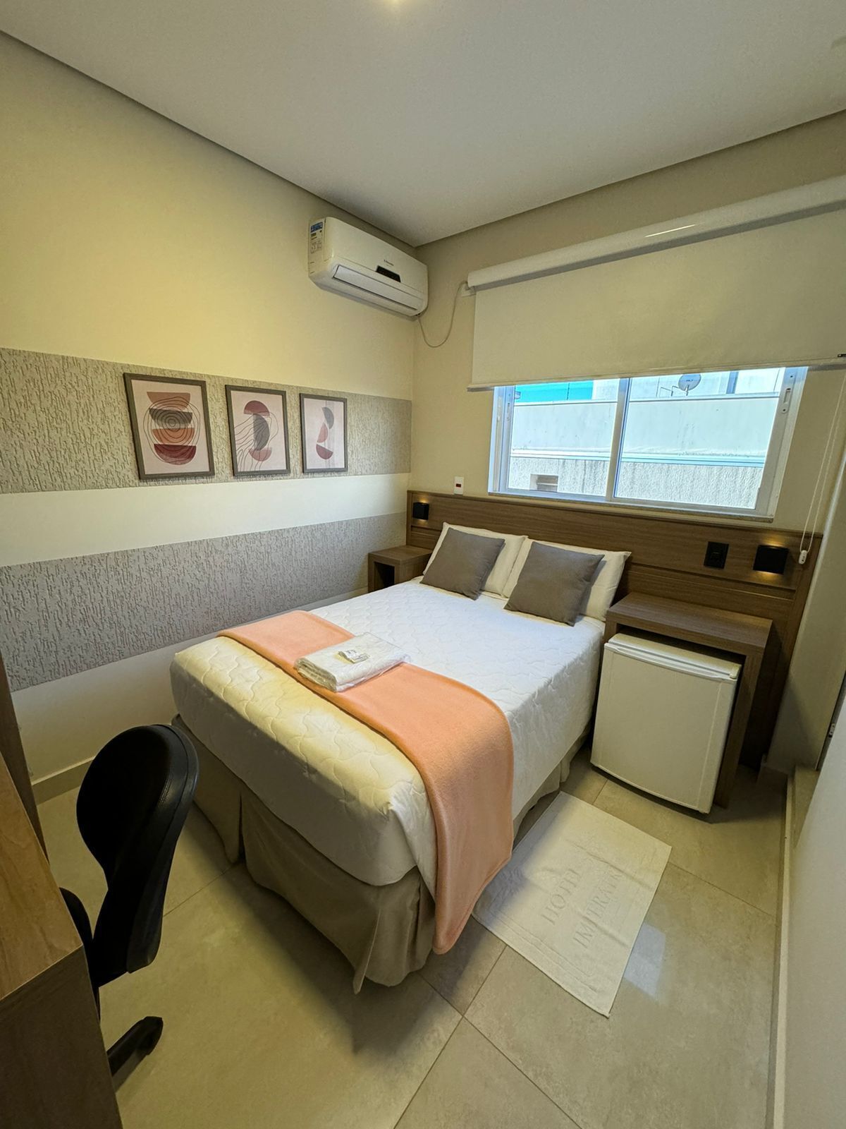 A bedroom with a bed, bedside tables, a desk, a chair, a small refrigerator, and artwork on the wall.