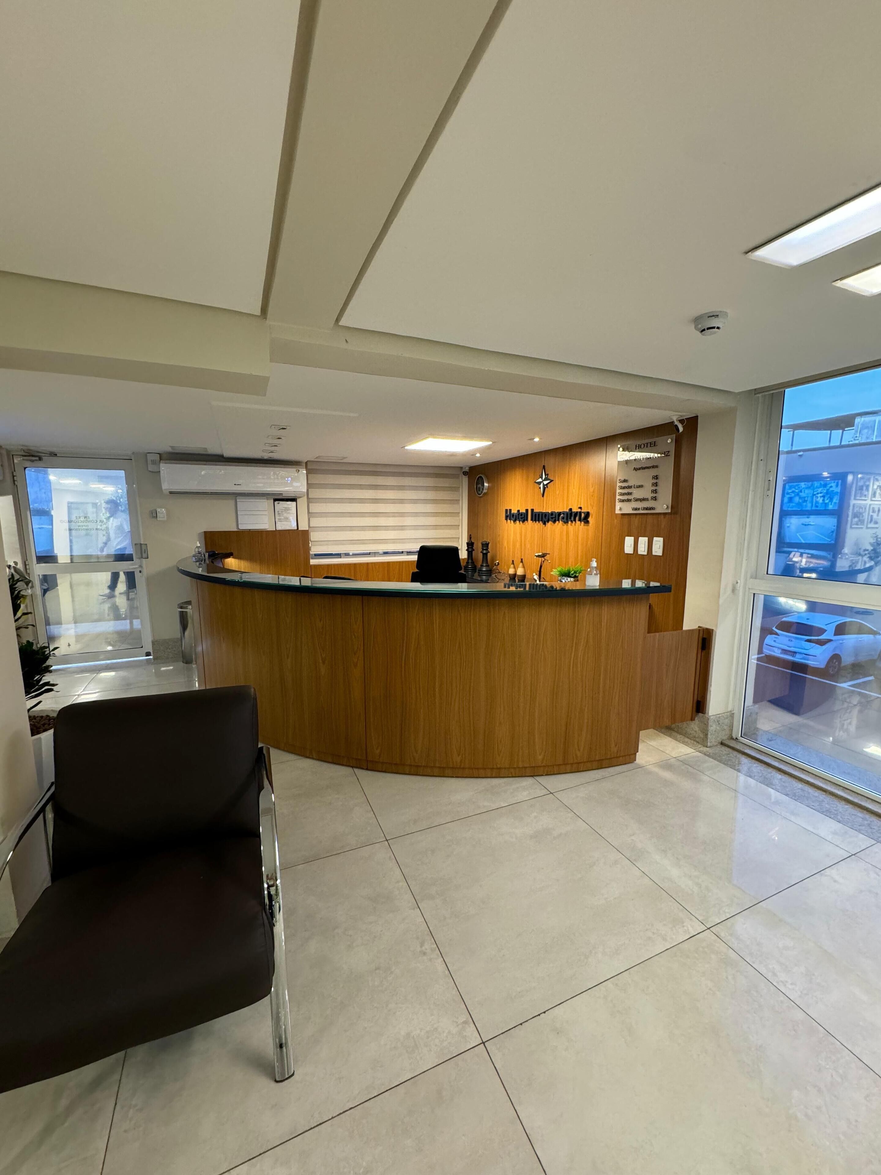 A hotel reception area with a wooden reception desk, a black chair, and a view of the parking area through a large window.