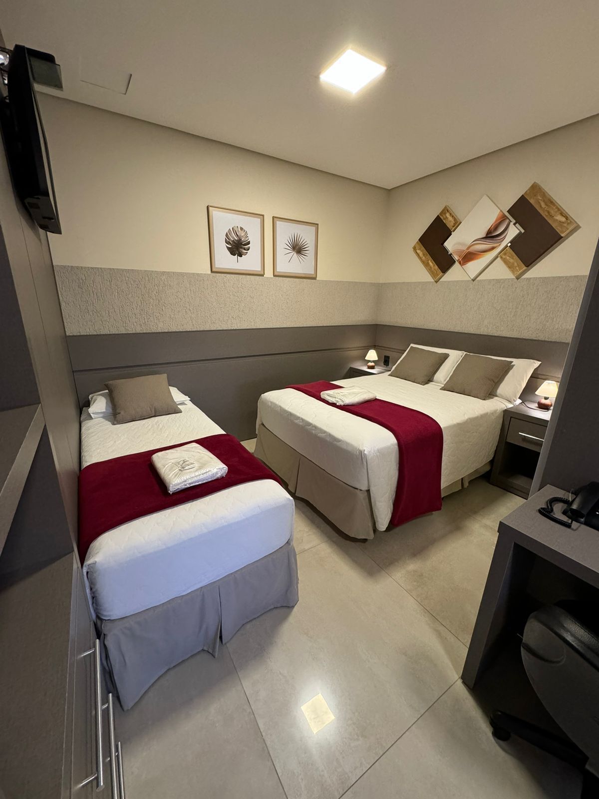 A hotel room with two beds, a desk, a chair, a TV, and artwork on the walls.