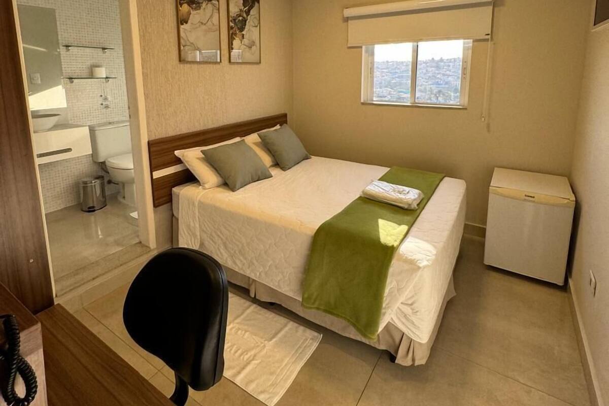 A hotel room with a bed, a desk, a chair, a small trash bin, and a window with a view.