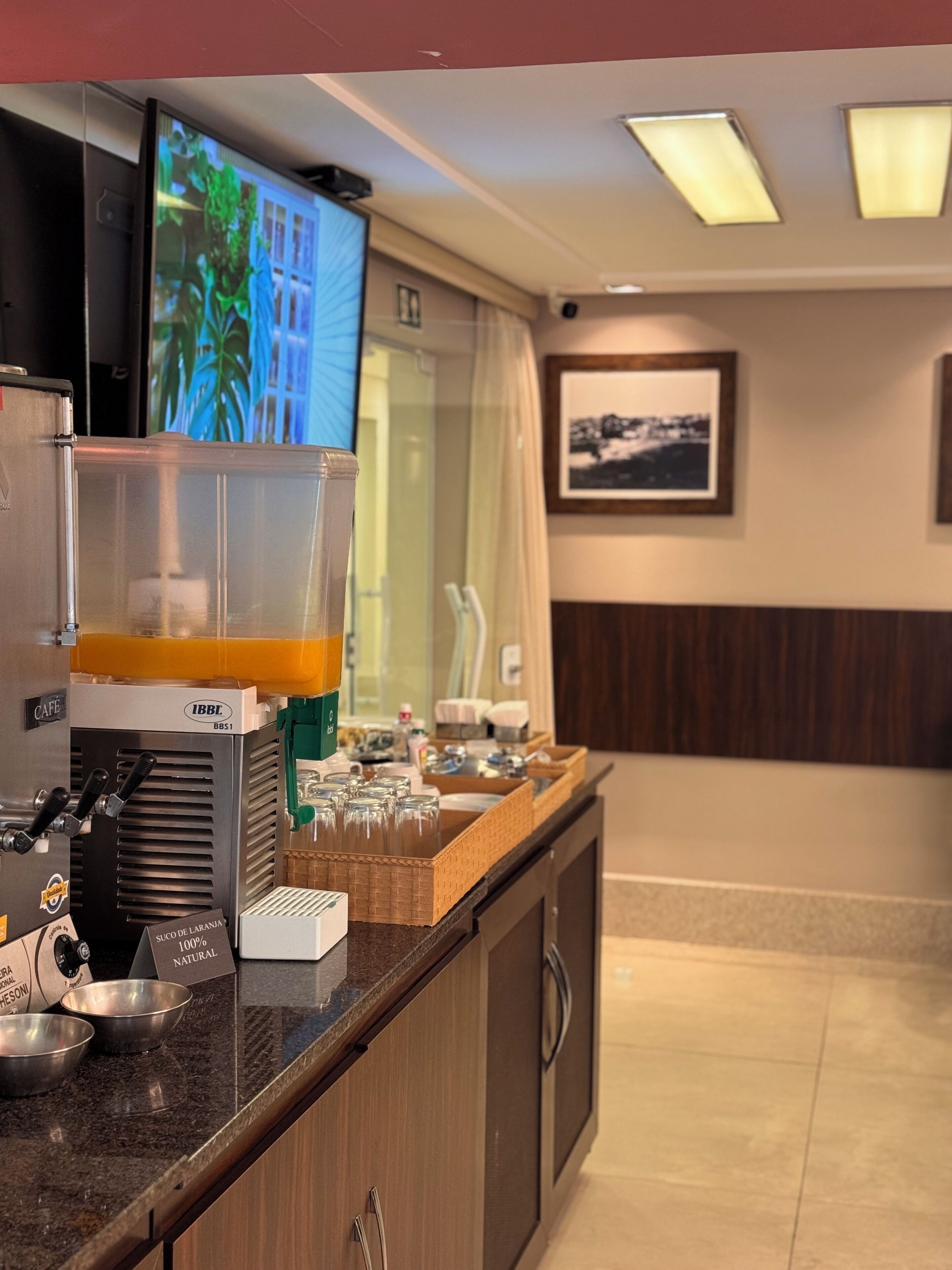 A coffee station with a coffee machine, a glass container, and a sign indicating the coffee is 100% natural. There is a television mounted on the wall displaying a nature scene.