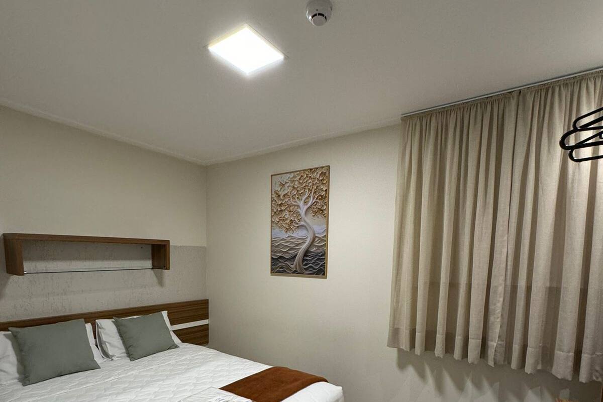 A hotel room with a bed, a headboard, a window with curtains, a wall-mounted artwork, and a desk with a chair.