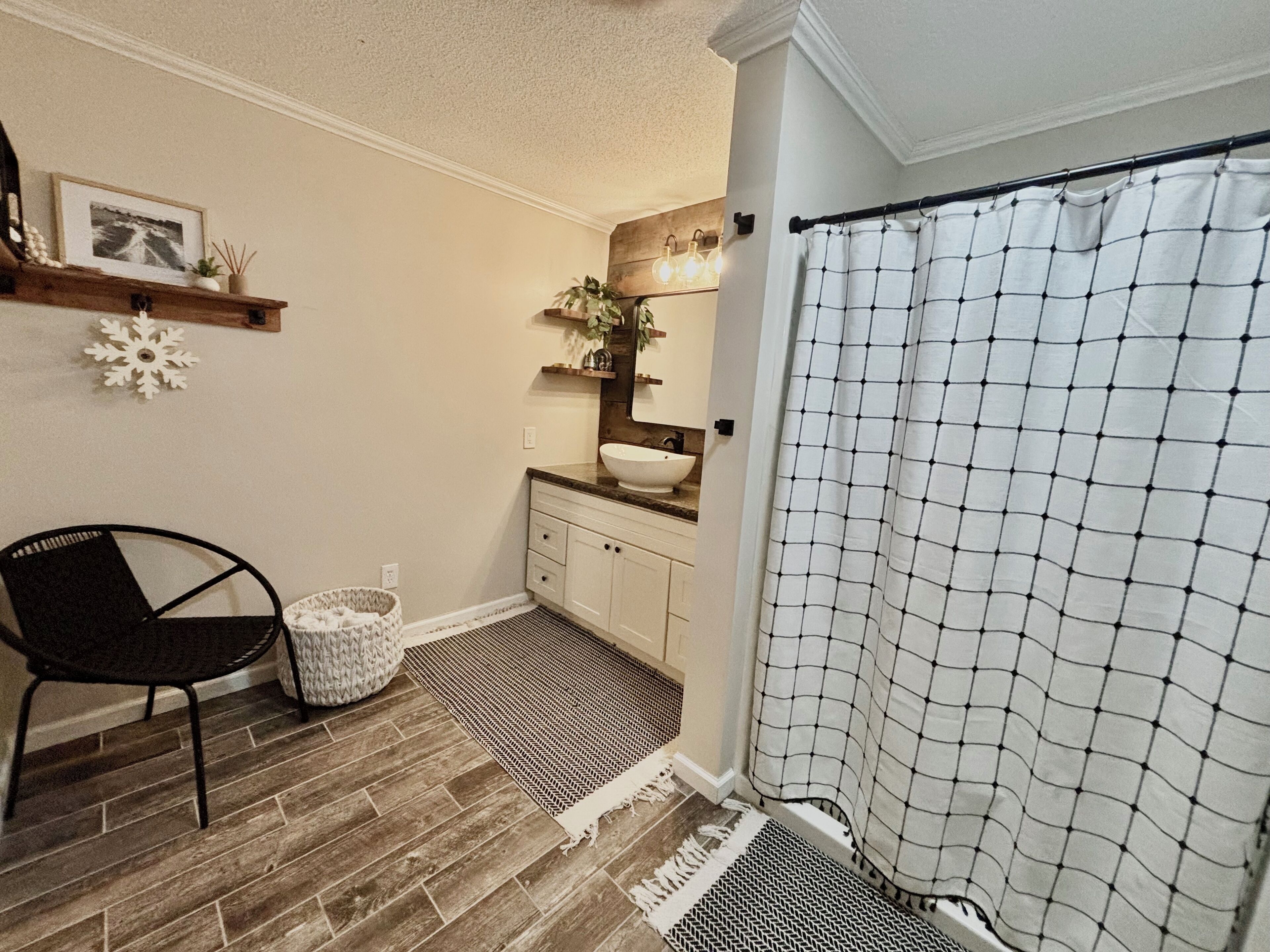 Une salle de bain avec un plancher en bois, une chaise noire, un lavabo blanc et un rideau de douche à motifs géométriques.