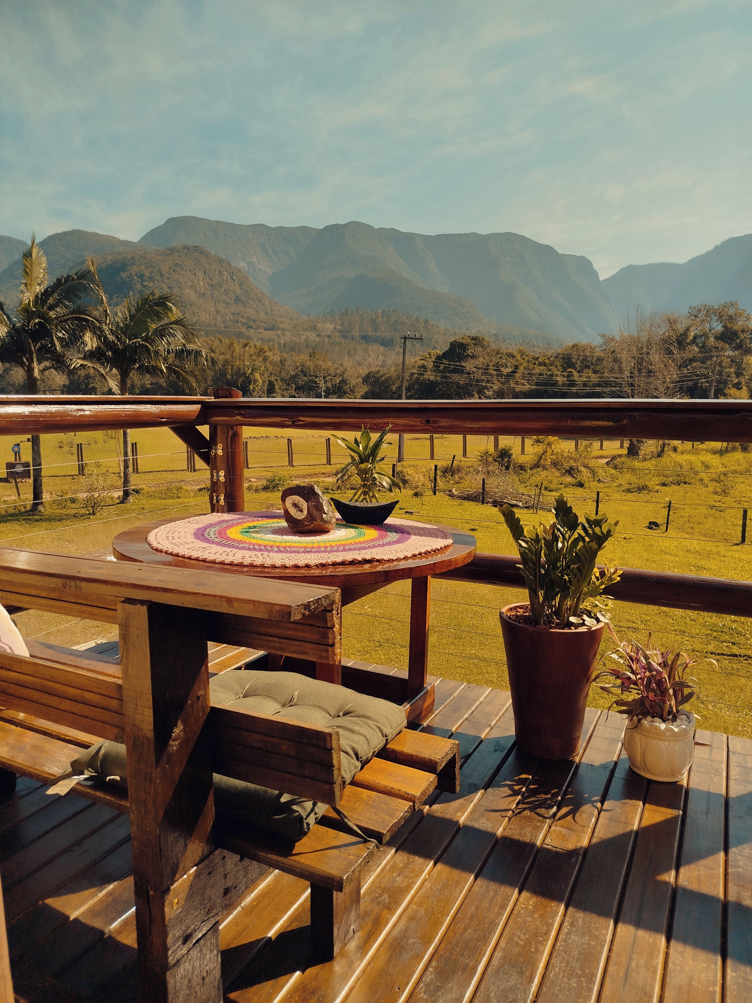 A wooden deck with a table and benches, overlooking a grassy field and mountains.
