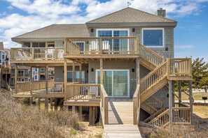 A two-story beachfront house with a wooden deck, a glass door entrance, and a staircase leading to the upper level.
