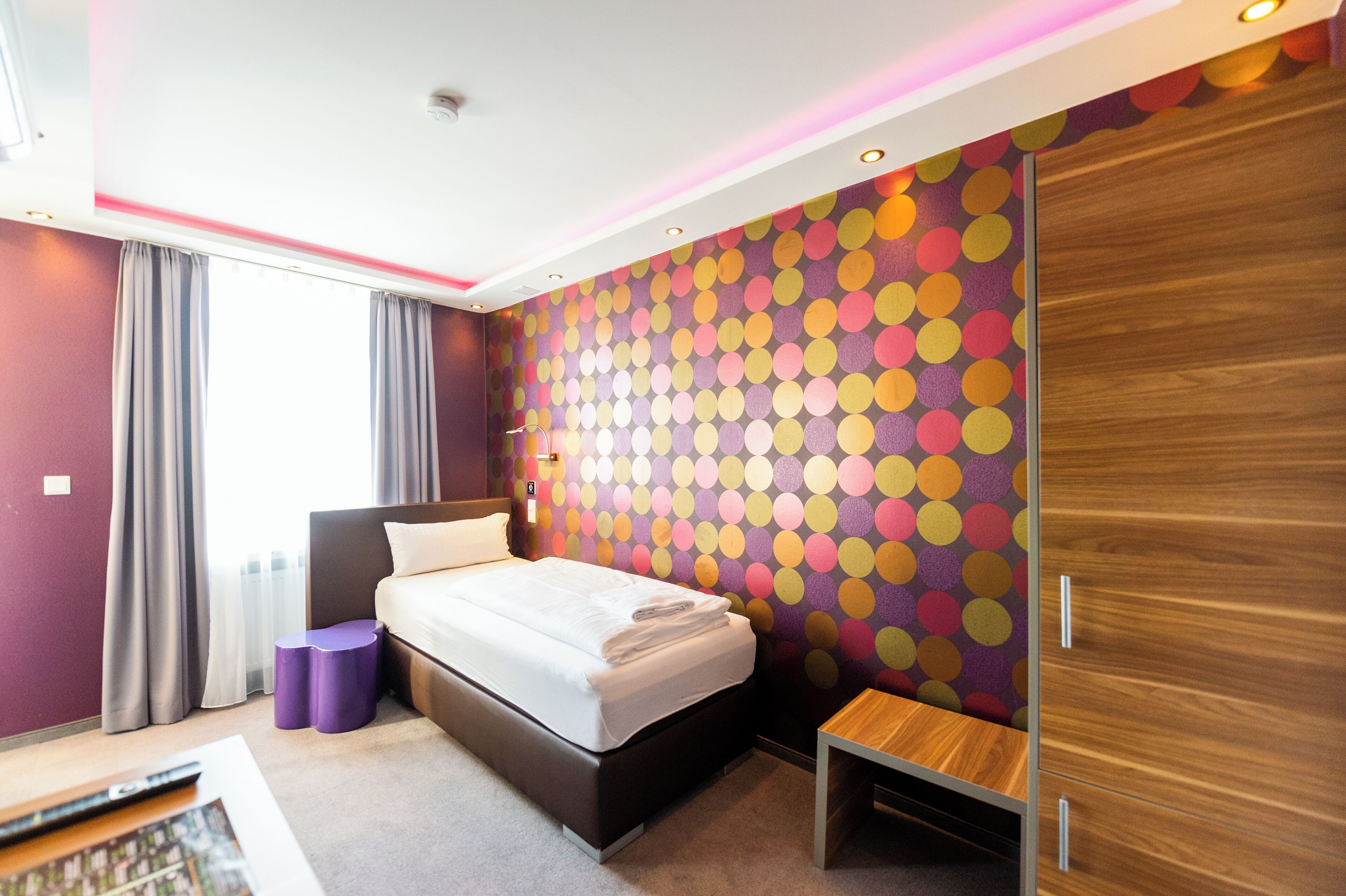 Ein modernes Hotelzimmer mit einem Bett, einem Holzschrank, einem kleinen Tisch und einer Wand mit bunten Punkten.