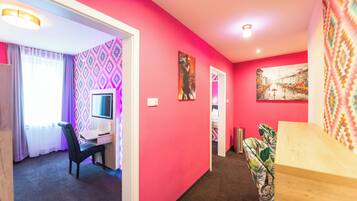 A room with a pink wall, a desk with a chair, a bed with a patterned bedspread, and a painting on the wall.