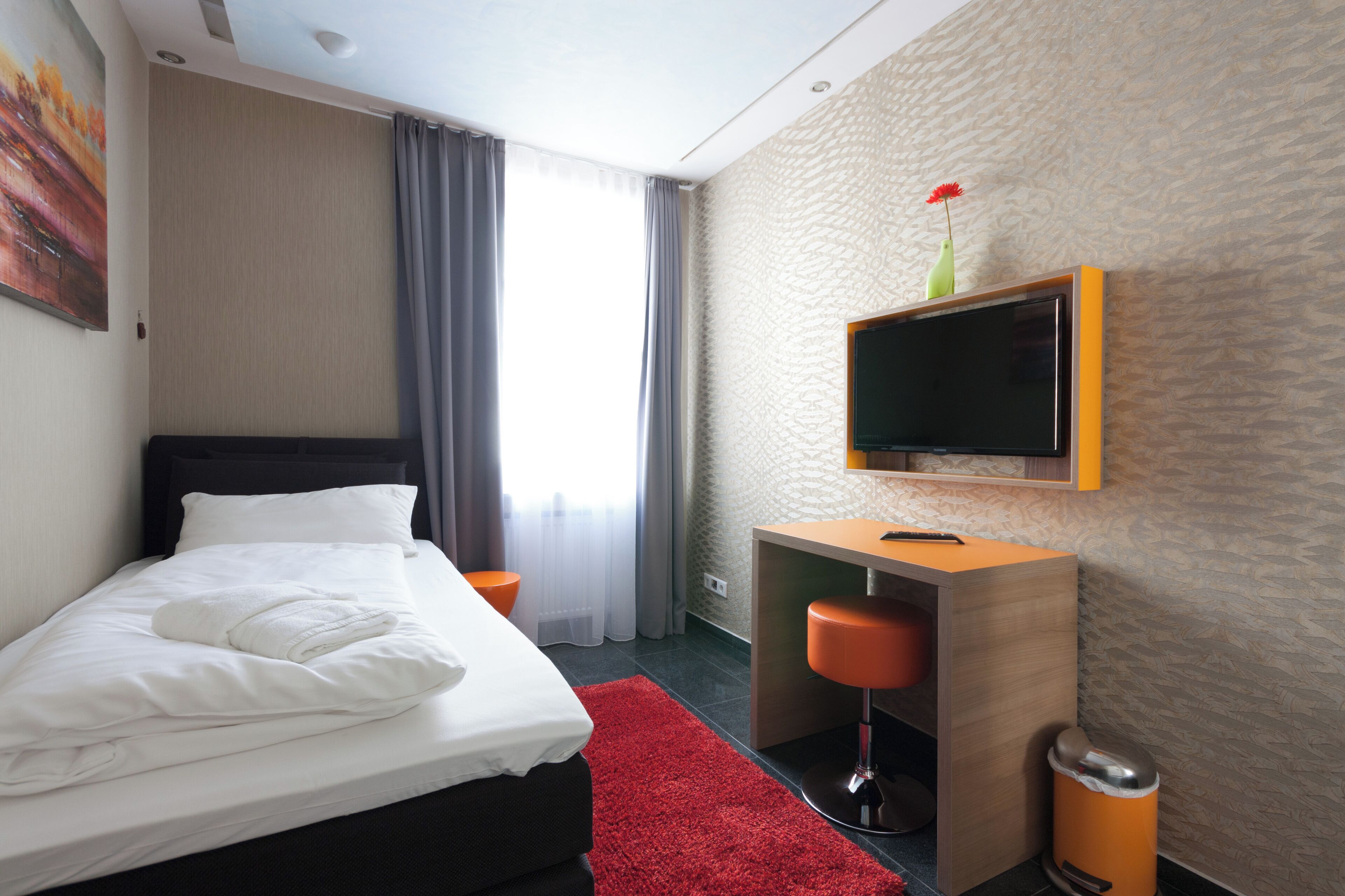 Habitación de hotel con cama, escritorio, silla, televisor montado en la pared y una alfombra roja.