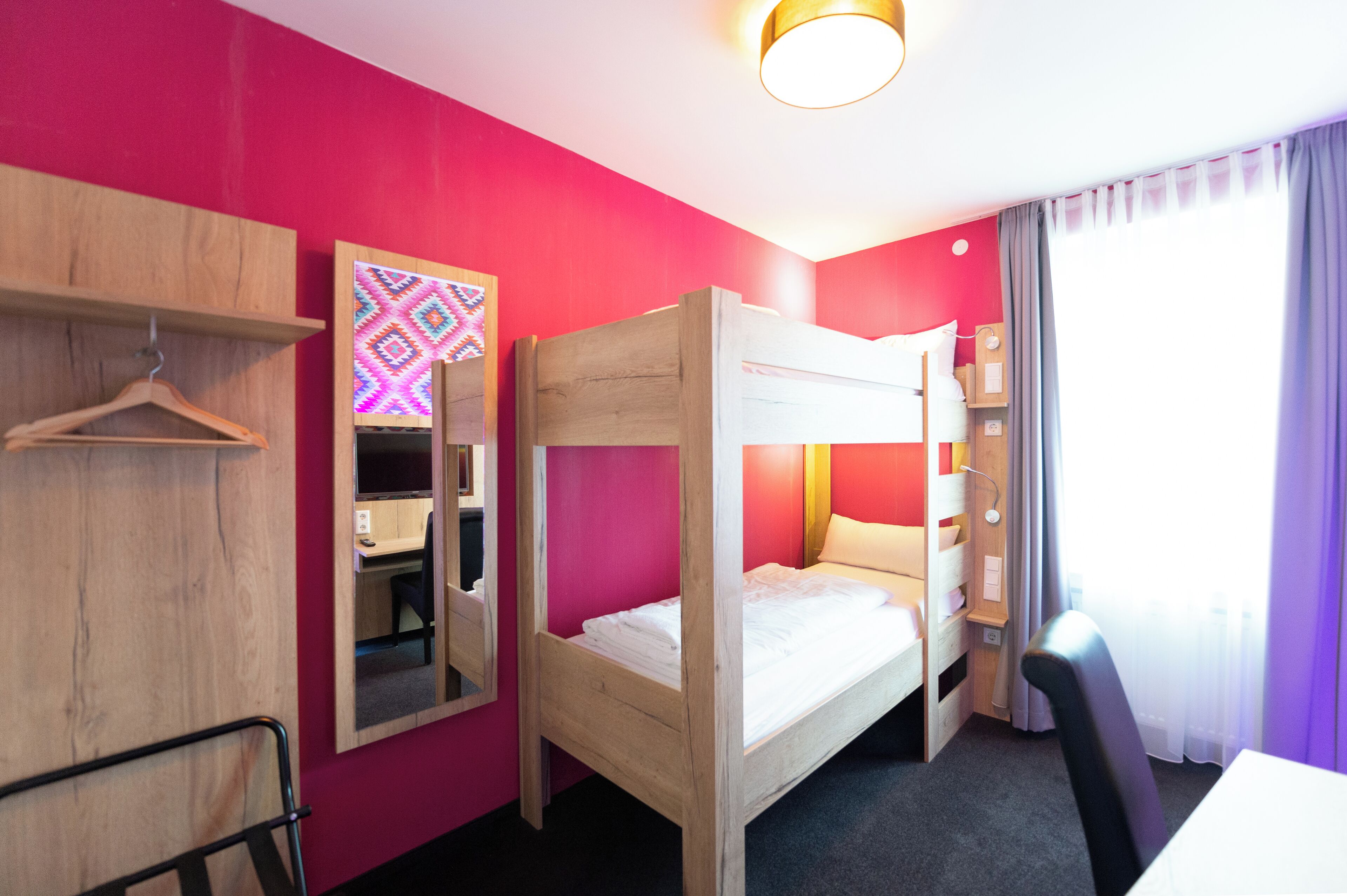 Een kamer met een stapelbed, een houten garderobe, een spiegel, een bureau en een stoel.