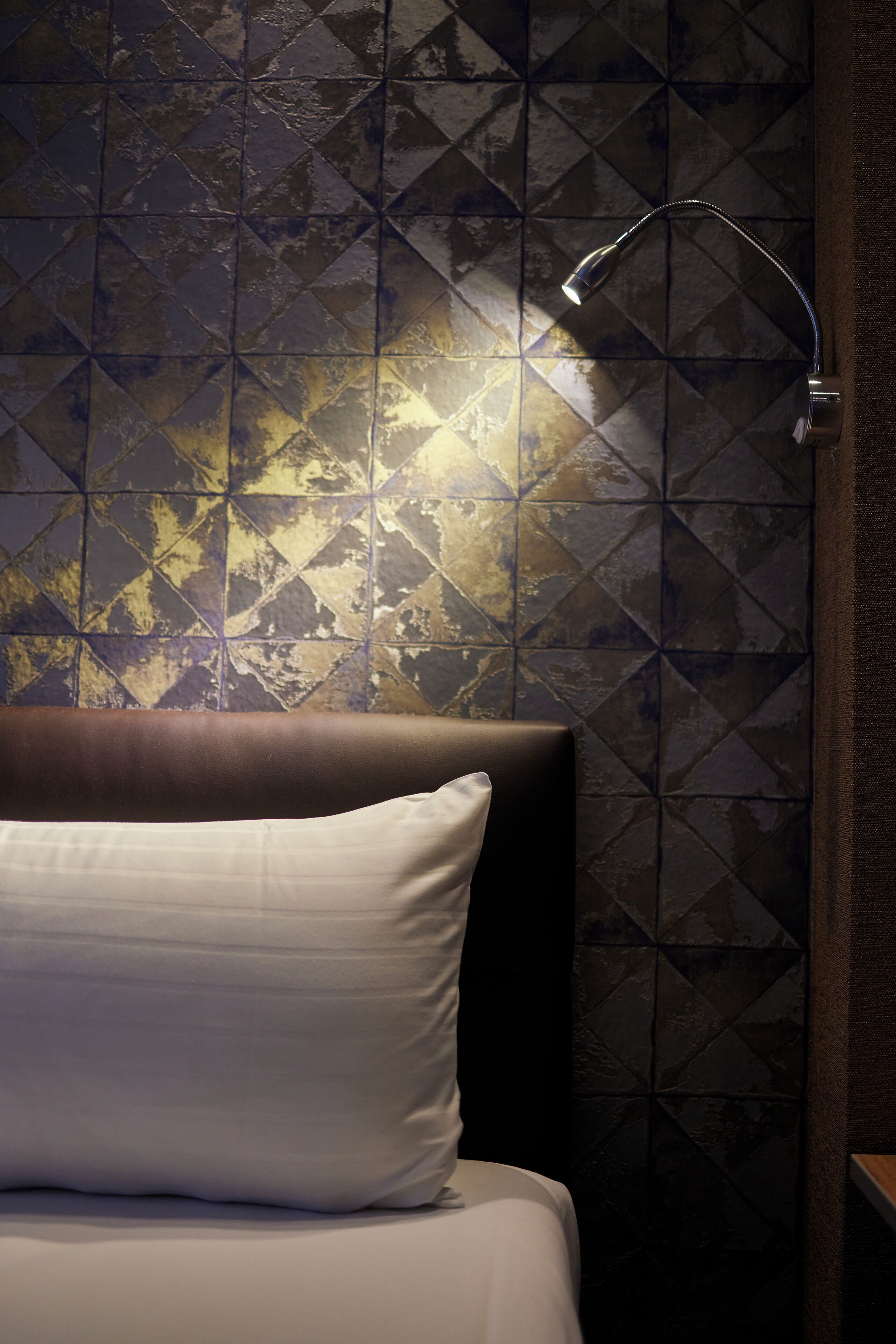 Una camera d'albergo con un letto, una testiera, un muro con motivo geometrico e un comodino con una lampada.
