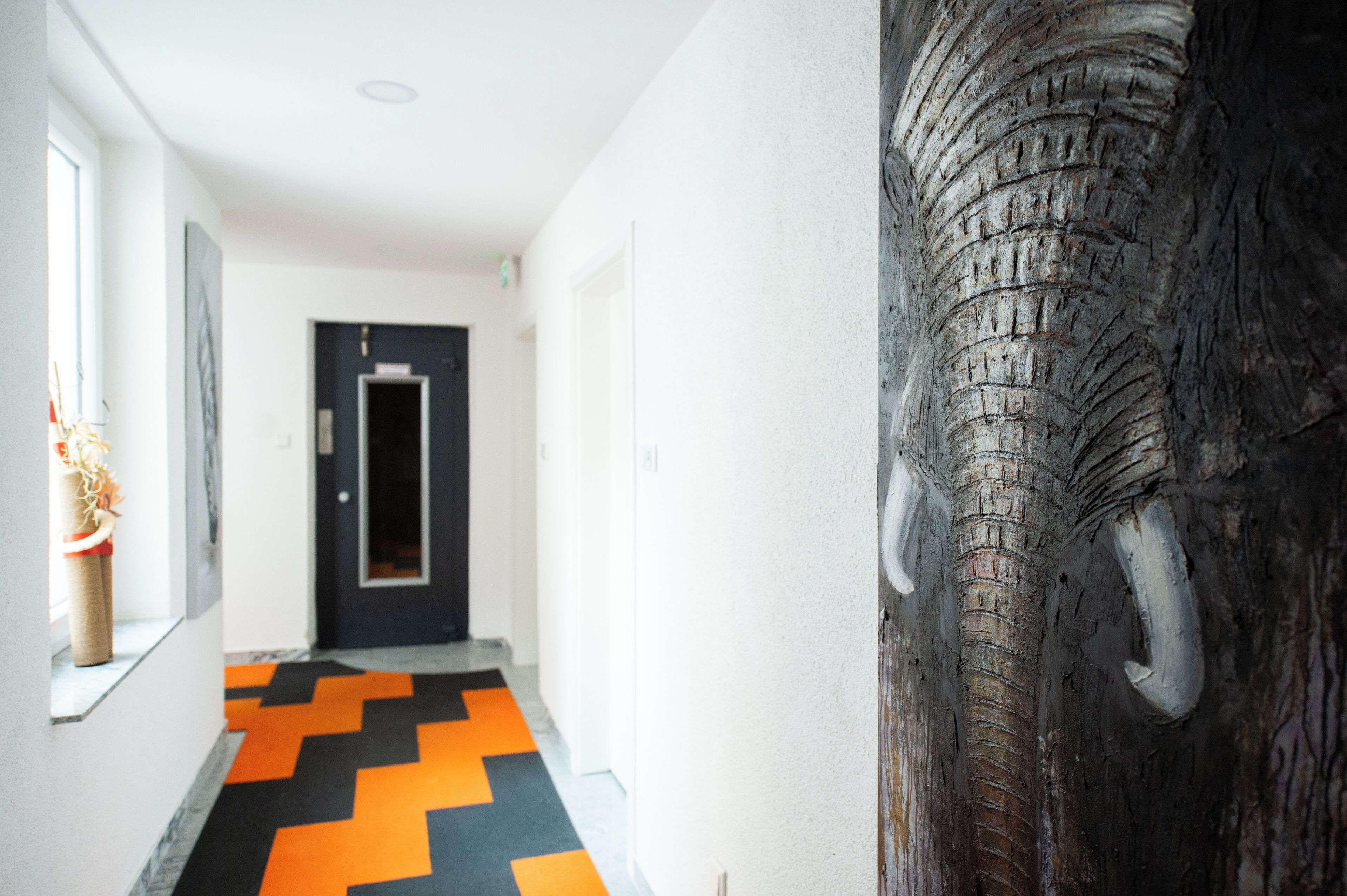 A hallway with a black and orange chevron-patterned floor, a door at the end, and a large elephant sculpture on the right wall.