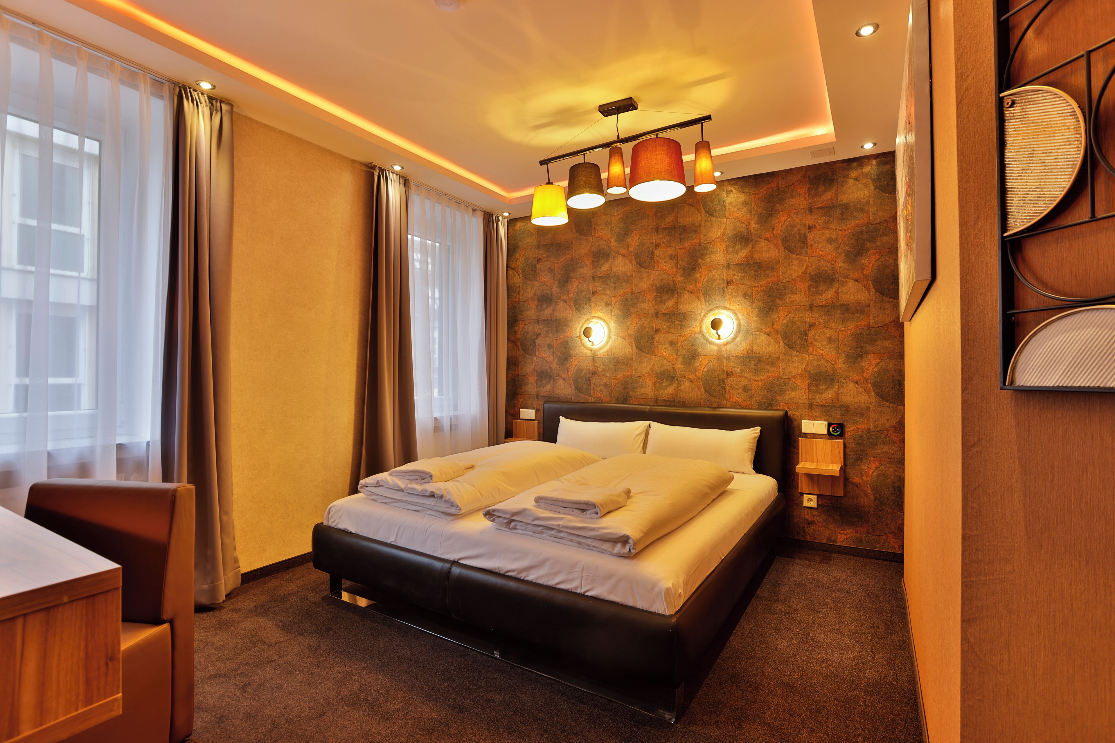 A hotel room with a large bed, a desk, and a chair. There are two wall-mounted lamps and a ceiling light fixture.