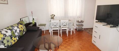 A compact living space with a sofa, a glass coffee table, a dining table with chairs, a TV, and a small cabinet.