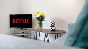 Een woonkamer met een televisie waarop 'NETFLIX' staat, een vaas met bloemen en een klein, rond tafeltje met een zwart beeldhouwwerk.