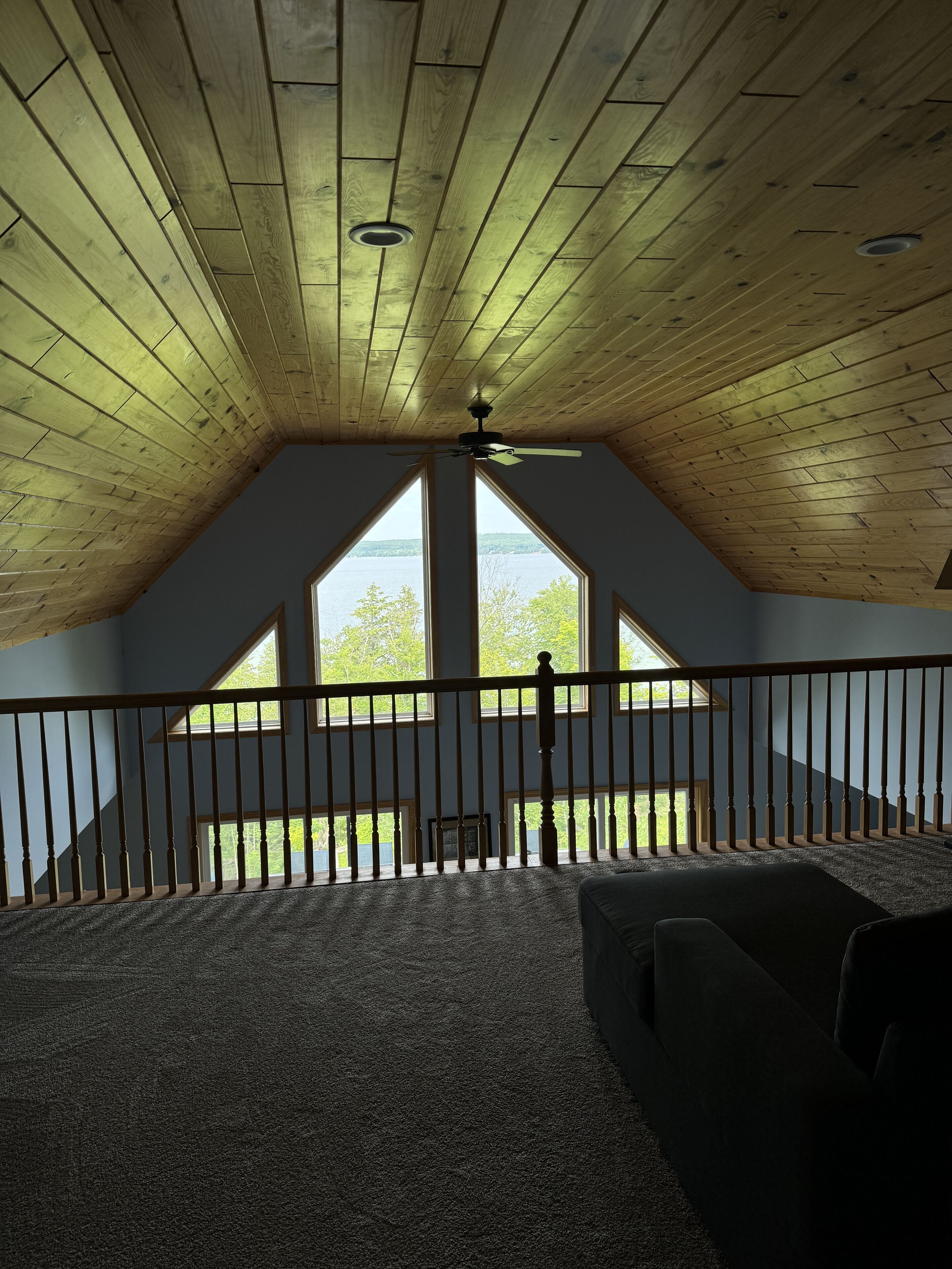 A room with a sloped ceiling, wooden beams, a ceiling fan, and a view of a lake through large windows.