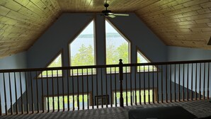 Ein Zimmer mit schrägem Dach, Holzbalken, Deckenventilator und Blick auf einen See durch große Fenster.