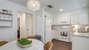 Una cocina moderna con armarios blancos, un microondas, un horno y un lavabo. Hay una mesa de comedor con mantel blanco y un cuenco con plantas verdes.