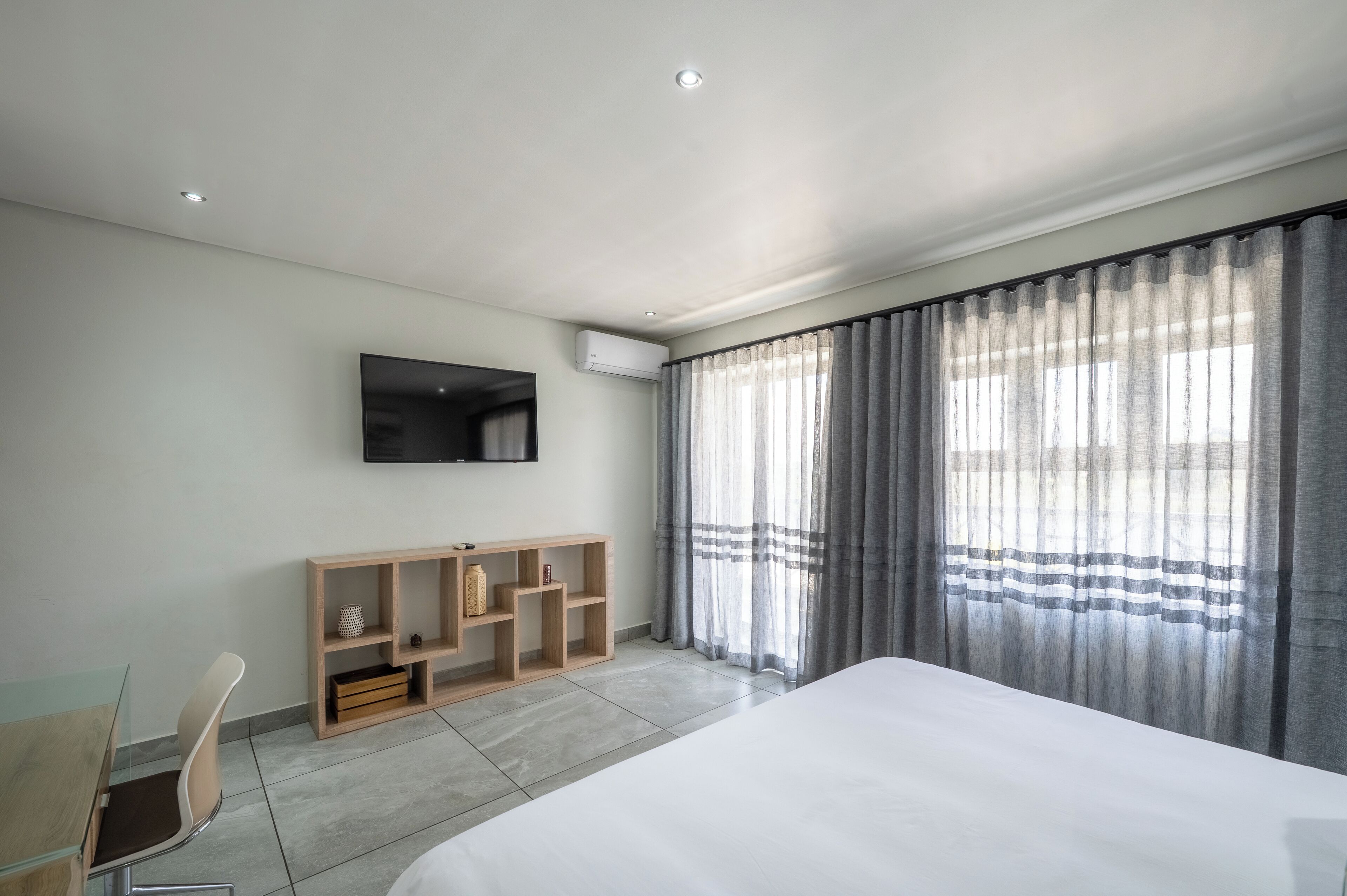 A modern hotel room with a large bed, a desk with a chair, a TV mounted on the wall, and a large window with curtains.