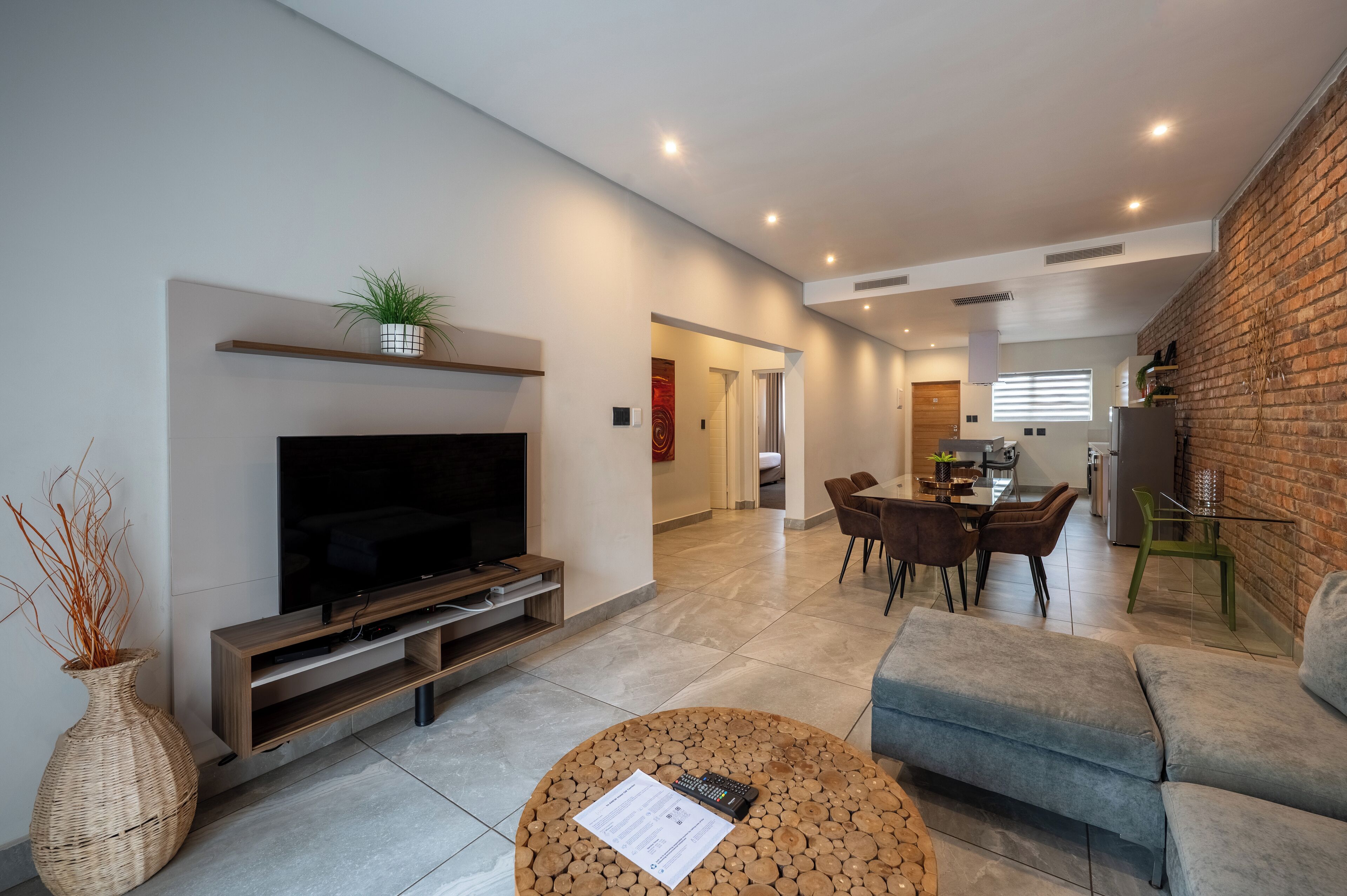 A modern living room with a flat-screen TV, a coffee table with a document, and a sofa. There is a dining area with a table and chairs, and a kitchen in the background.