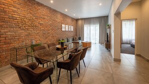 A modern living room with a glass dining table, brown chairs, a sofa, and a brick wall. - ANEW Hotel & Convention Centre OR Tambo (Benoni)