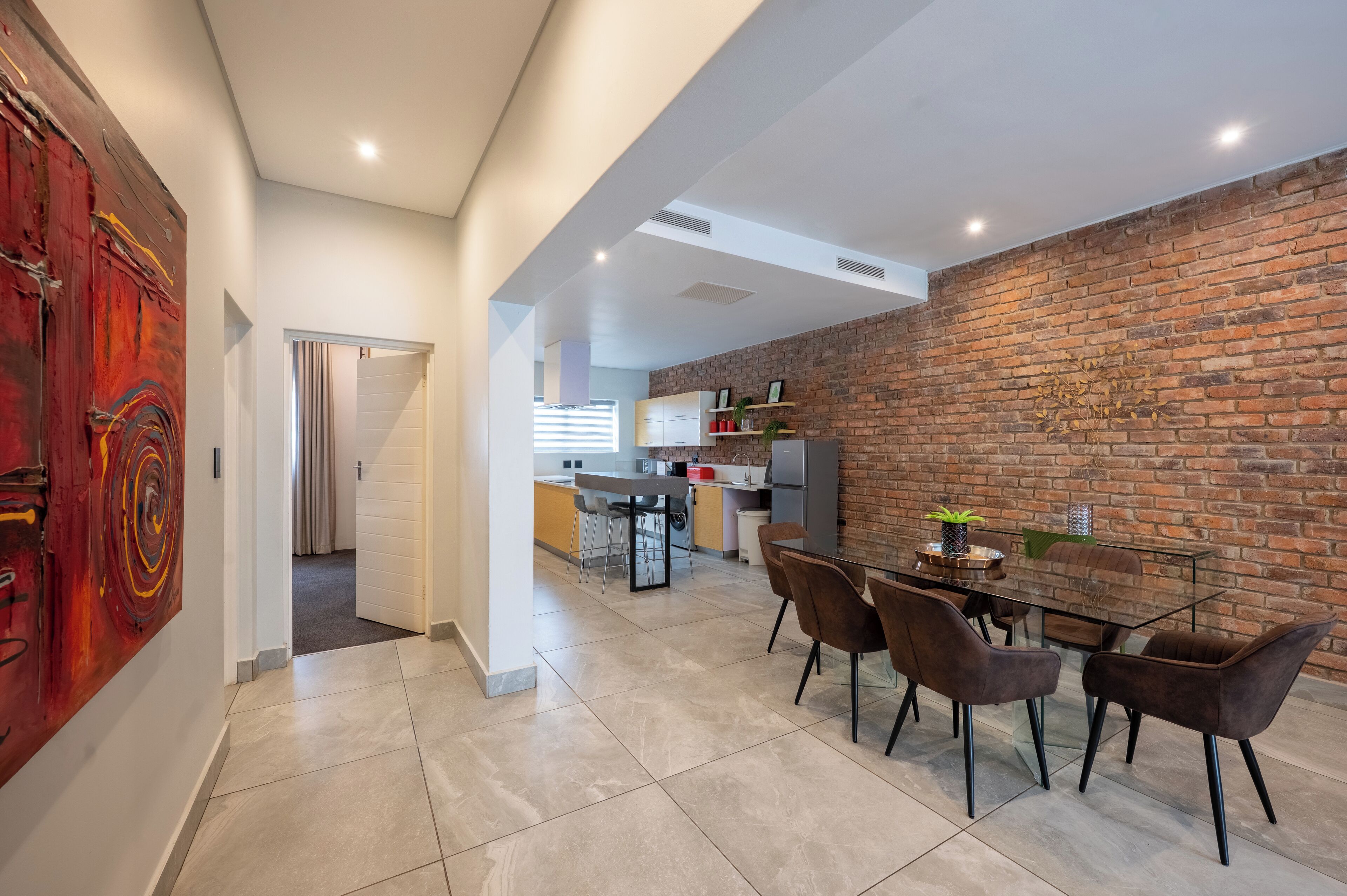 A modern kitchen with a brick wall, a dining area with a glass table, and a red abstract painting.
