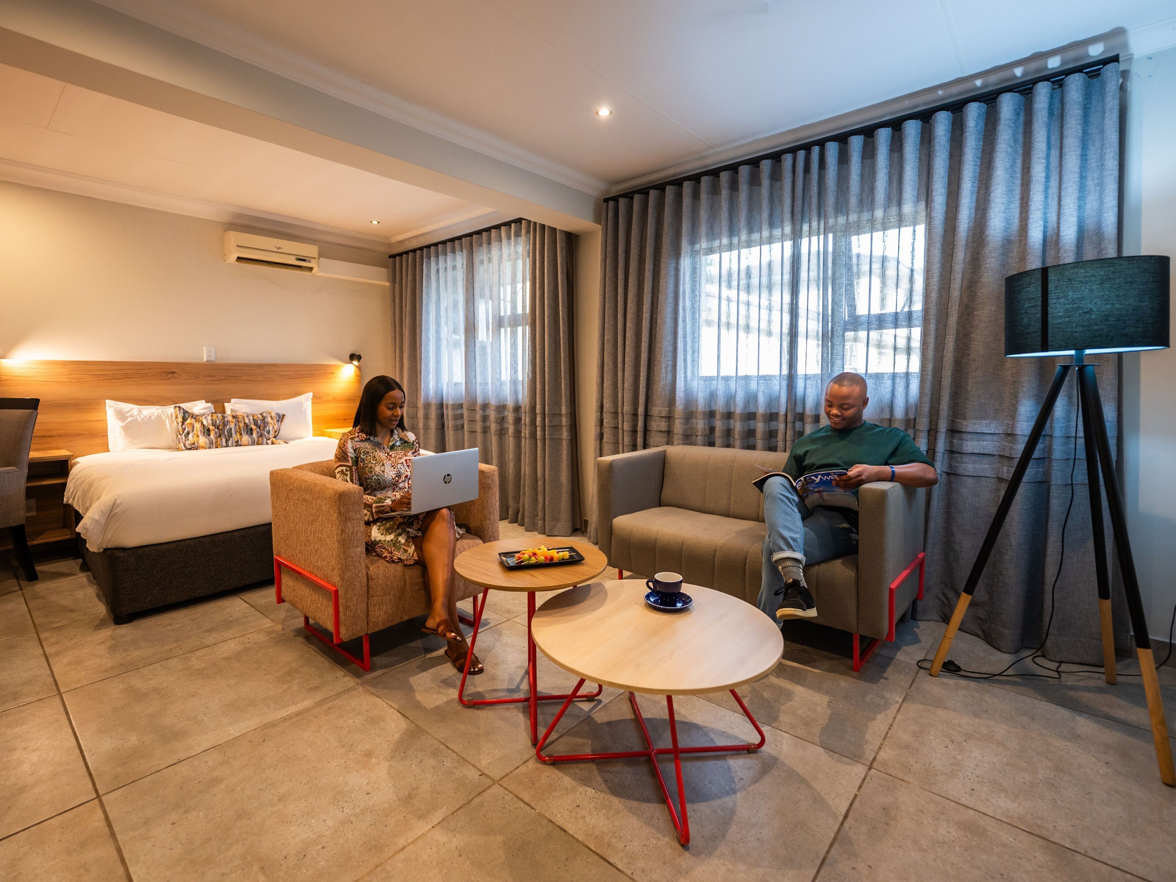 A hotel room with a bed, two armchairs, a small table, a tripod, and curtains.