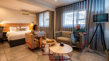A hotel room with a bed, two armchairs, a small table, a tripod, and curtains.