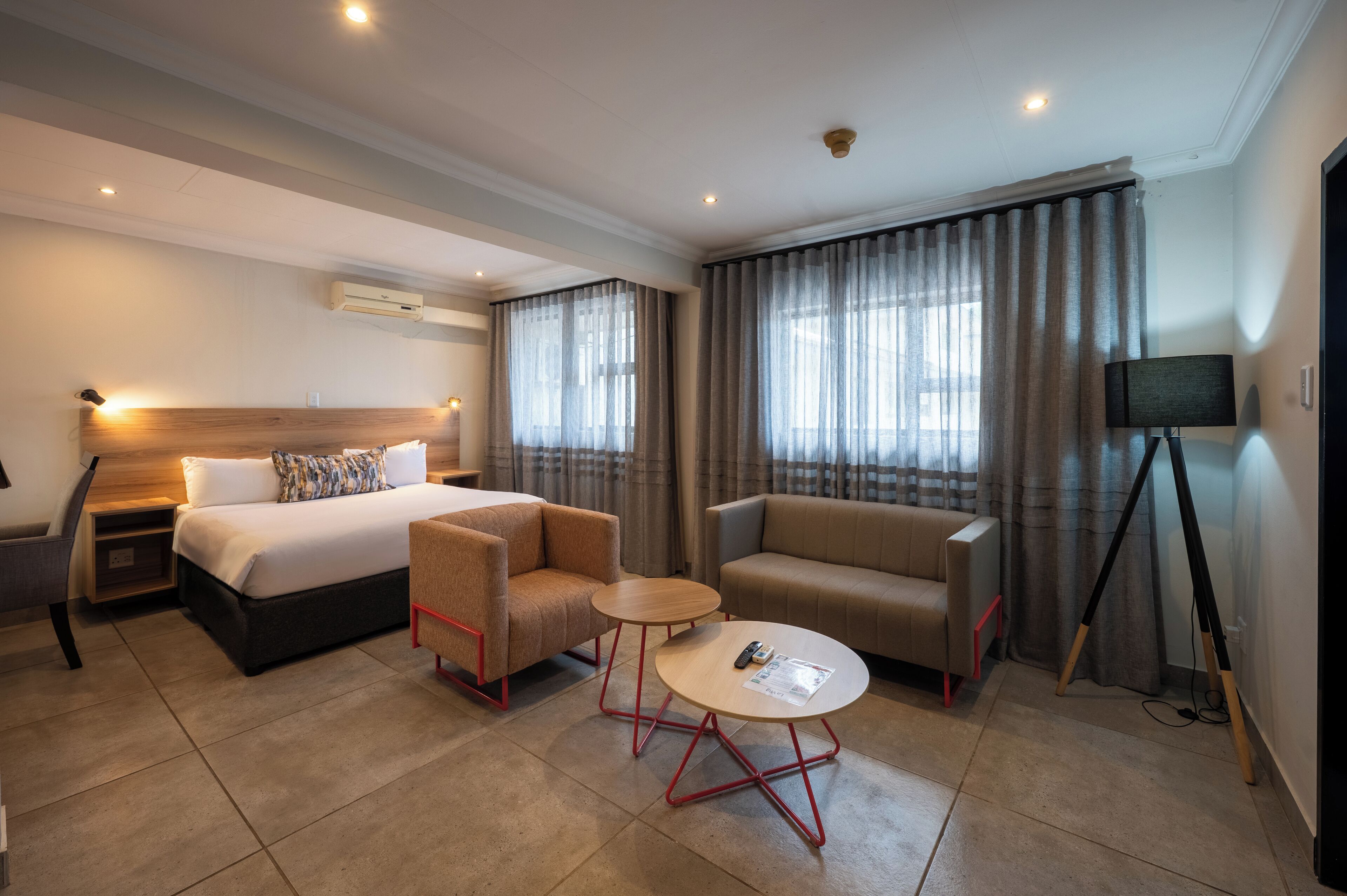 A hotel room with a bed, two armchairs, a small table, a TV on a tripod, and curtains.