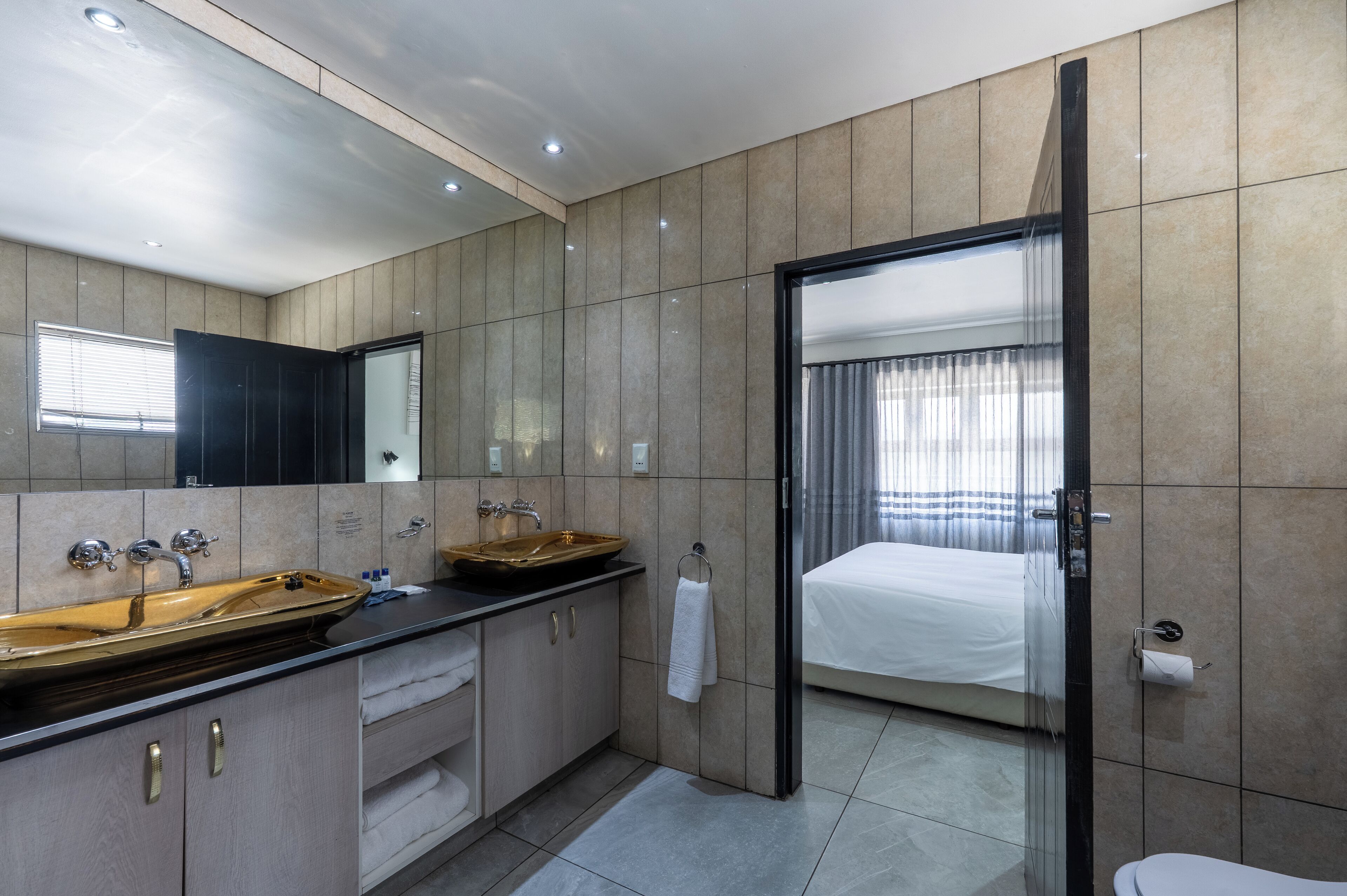 A modern bathroom with a double sink, a large mirror, and a glass door leading to a bedroom.