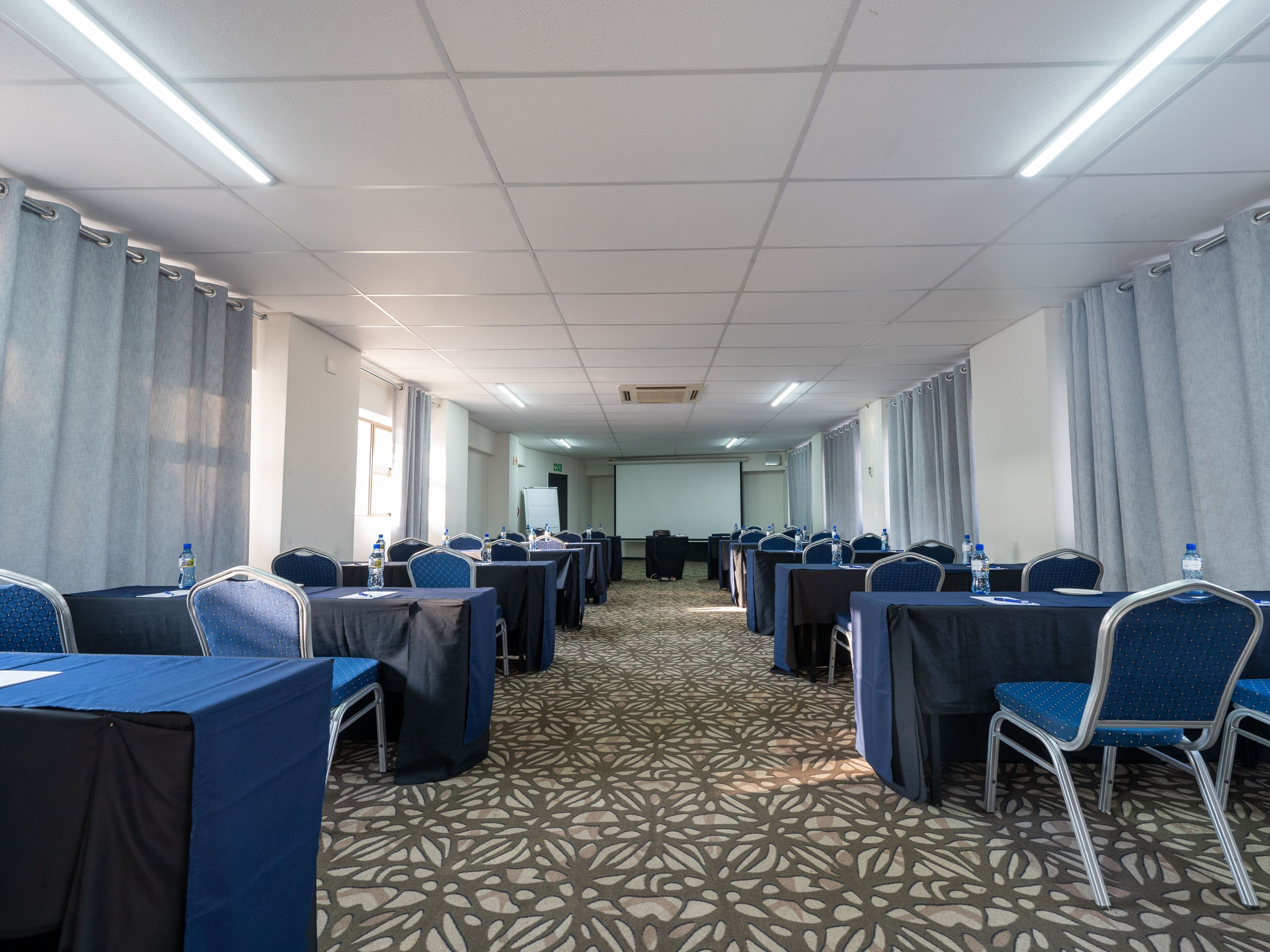 Une salle de conférence avec des rangées de tables et de chaises, une moquette à motifs et un écran de projection à l’avant.
