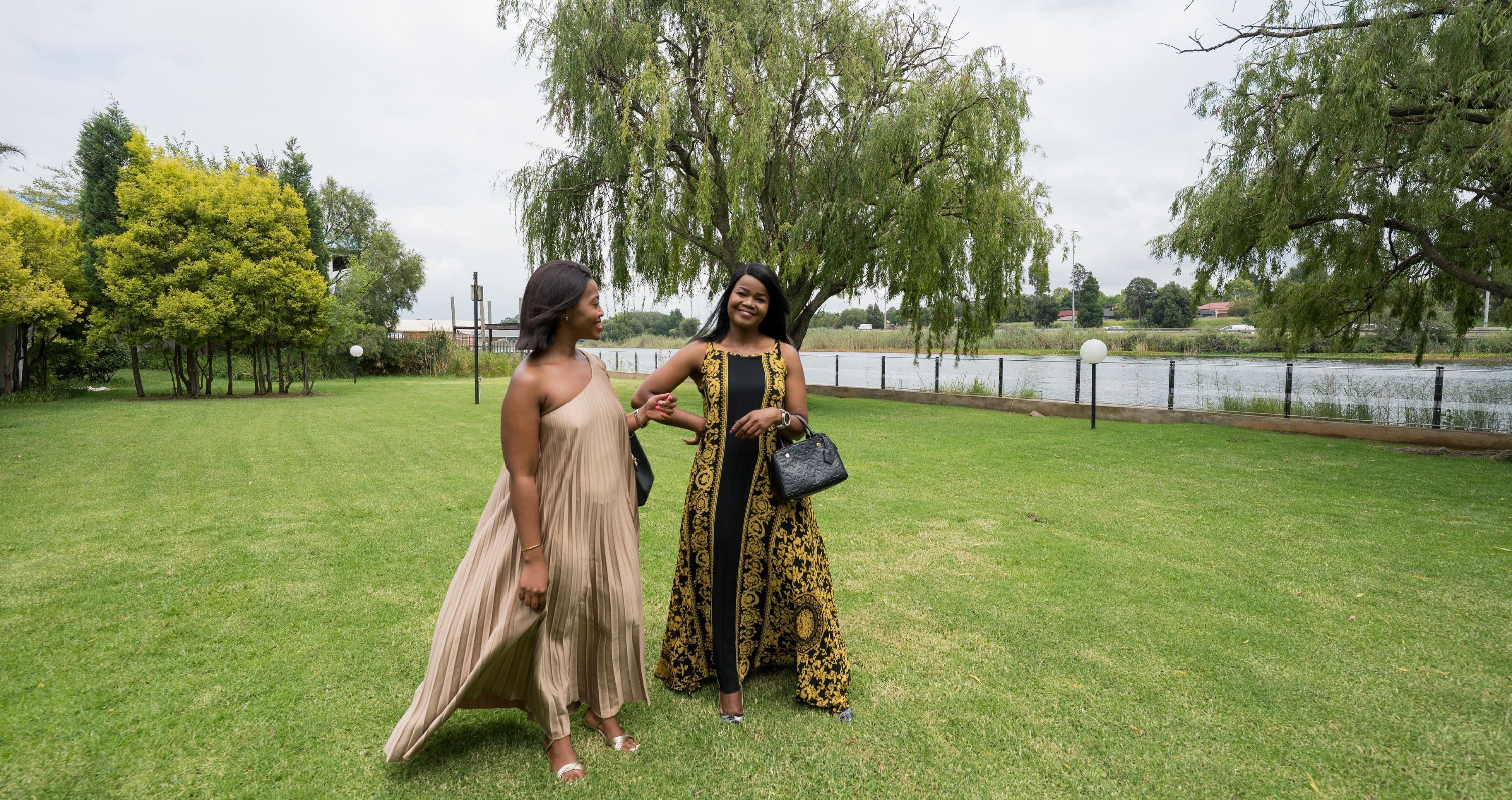 Two women walking on a grassy area with trees and a body of water in the background.