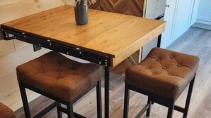 A wooden dining table with two cushioned stools, a vase with dried plants, and a kitchen area with white cabinets and a toaster.