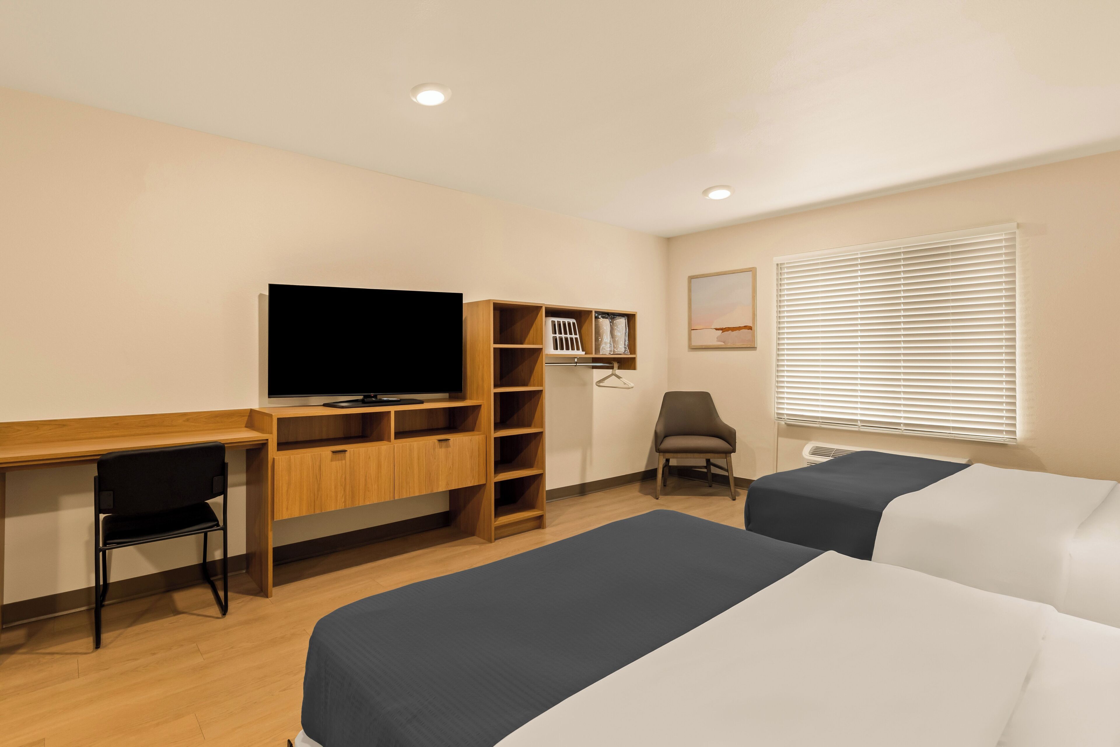 Habitación de hotel con escritorio de madera, televisor, una silla, dos camas, armario y ventana con persianas.