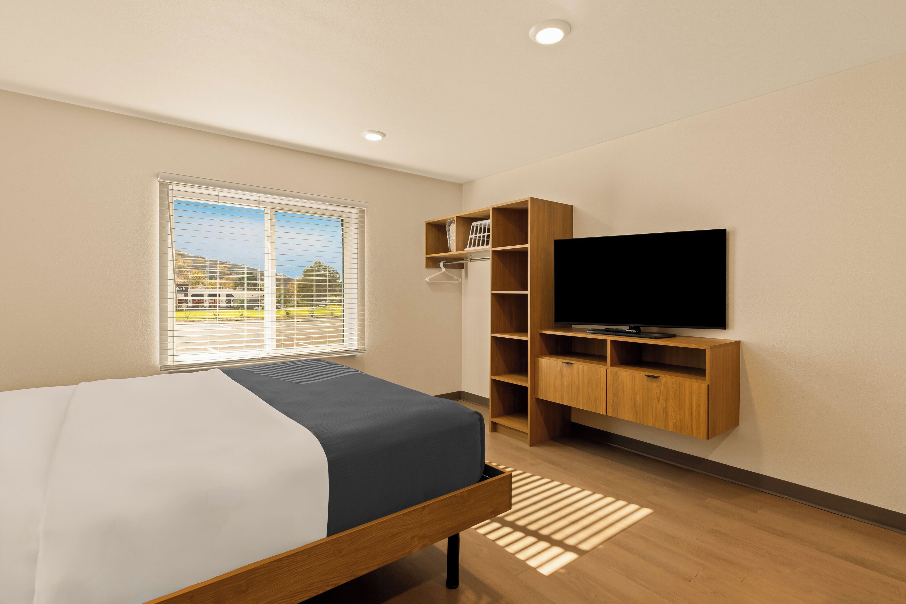 Une chambre d’hôtel moderne équipée d’un lit, d’un téléviseur, d’un meuble en bois pour le téléviseur, d’une fenêtre avec des stores et donnant sur l’extérieur.