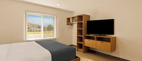 Una habitación de hotel moderna con una cama, un televisor, un mueble de televisión de madera, una ventana con persianas y vistas al exterior.