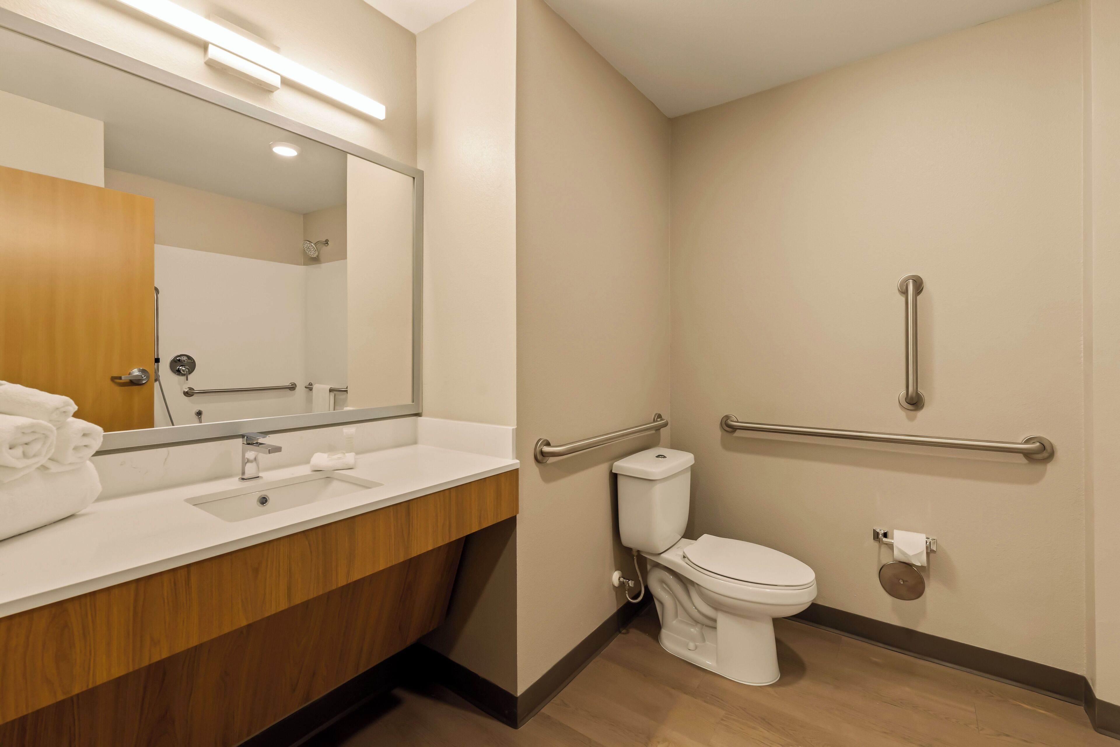 A modern bathroom with a wooden vanity, a large mirror, a toilet with a handrail, and a shower area.