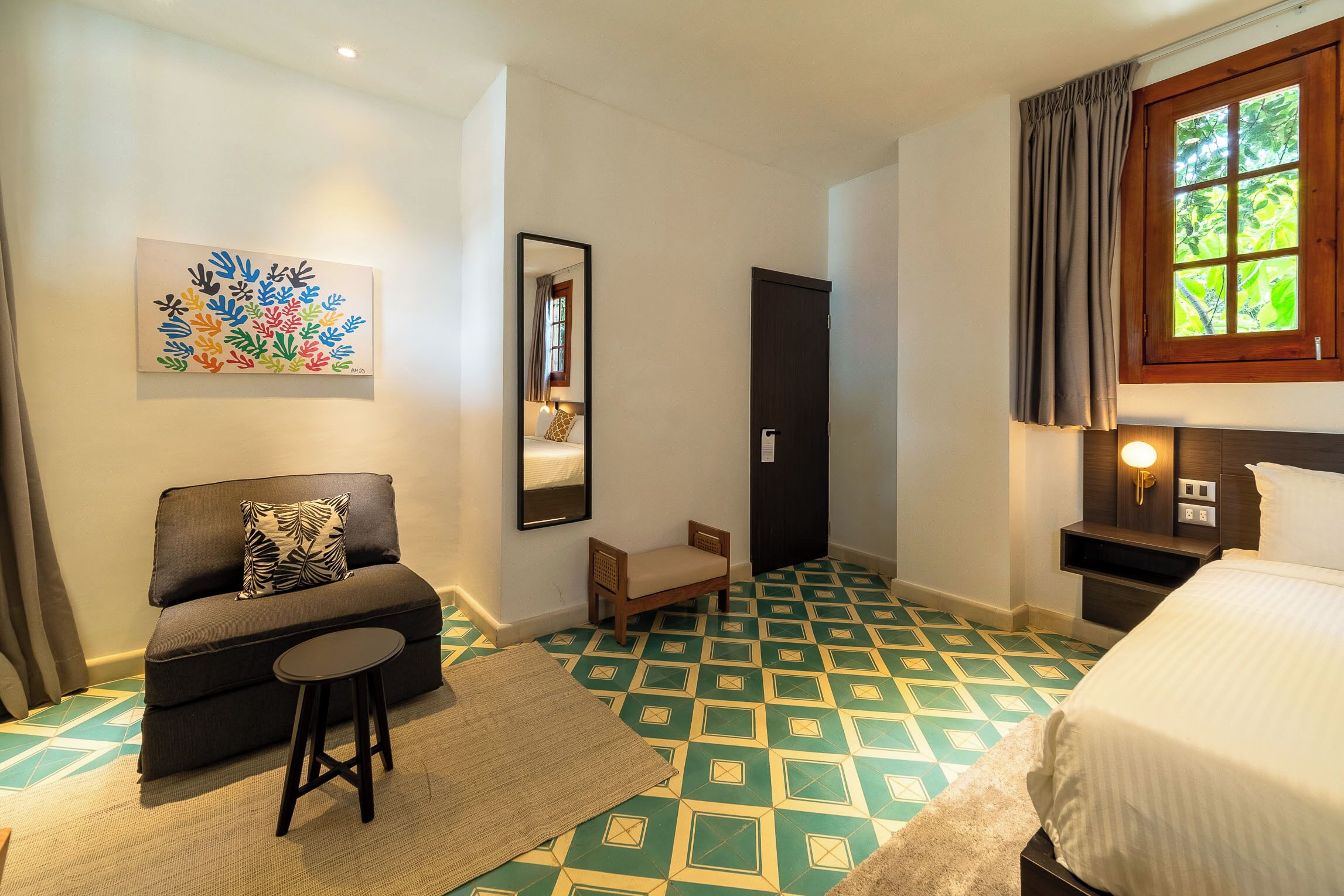 Quarto de hotel com sofá, um banquinho, cama, mesa de cabeceira, espelho e um padrão de cores no chão.