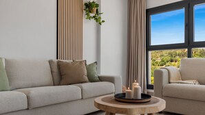 Una sala moderna con un sofá beige, una mesa de centro redonda de madera con velas, y vistas a un paisaje verde a través de grandes ventanales.