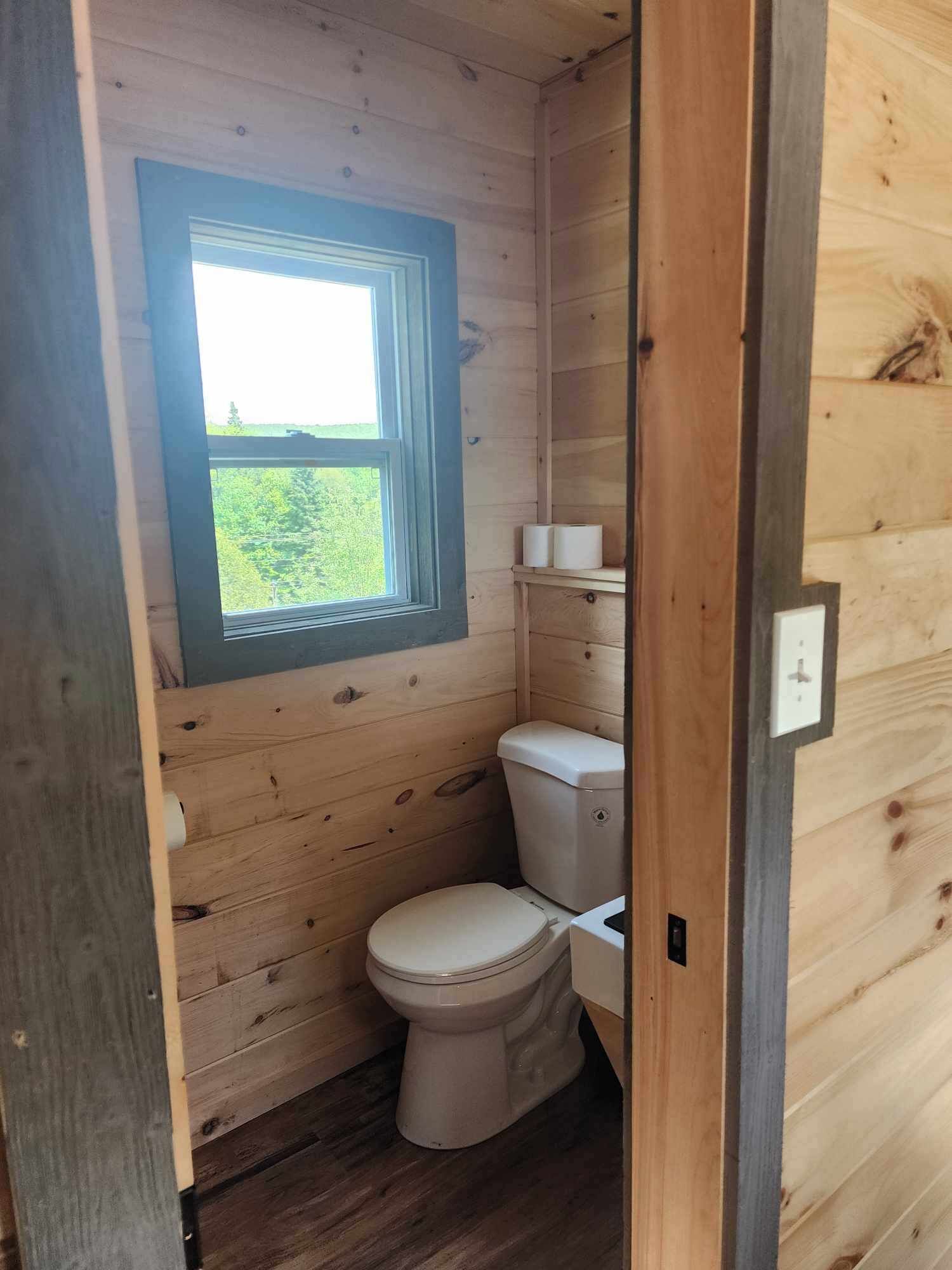 A small, wooden-paneled bathroom with a toilet, a window, and a shelf with toiletries.