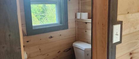 A small, wooden-paneled bathroom with a toilet, a window, and a shelf with toiletries.